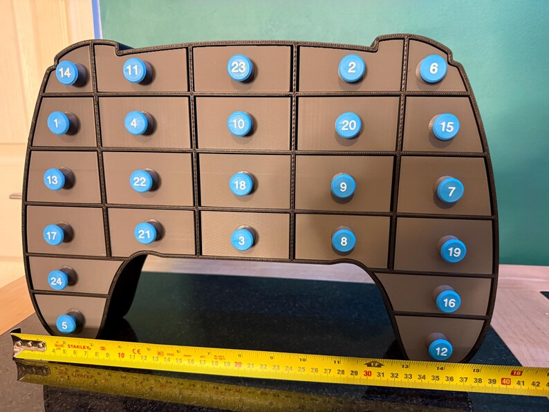 May include: A brown, multi-compartment advent calendar with blue knobs, numbered 1-24. The calendar is shaped like a rounded rectangle. A yellow tape measure shows a length of approximately 16 inches.