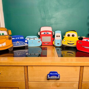May include: A collection of colorful, cartoon-style car cutouts, including a yellow taxi, a red race car, and various other car designs. The cars are arranged on a wooden surface, with a blue car-shaped drawer pull in the foreground.