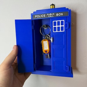 May include: A blue key holder shaped like a police call box with the words "POLICE PUBLIC CALL BOX" on top. The door is open, revealing keys with a tag. The key holder is mounted on a white wall.
