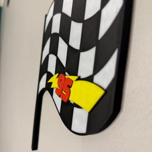May include: A black and white checkered flag with a red "95" and yellow lightning bolt design. The flag has a black handle and is likely a decorative item or a party decoration.