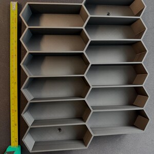May include: A dark gray modular wall organizer featuring a honeycomb pattern with ten hexagonal compartments. A yellow measuring tape is positioned along the left side, indicating the total height is approximately 10.5 inches.