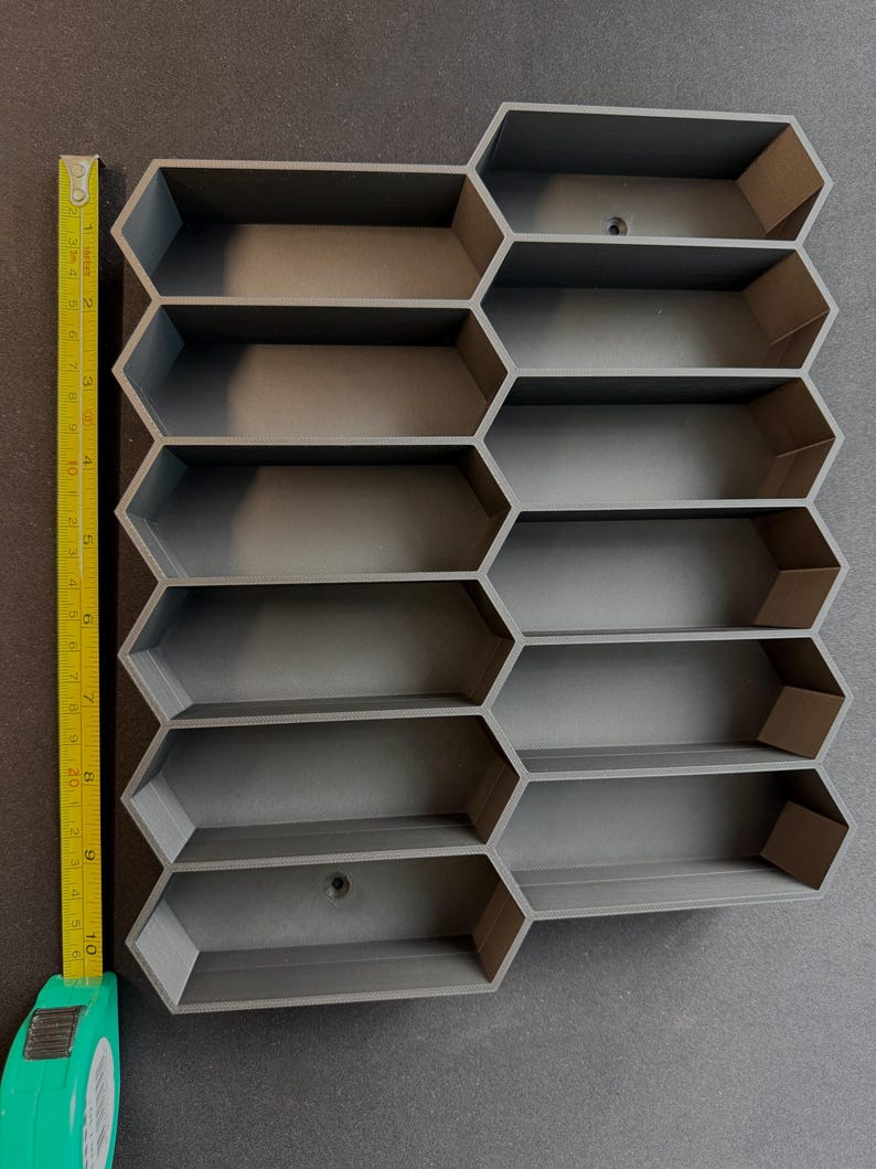 May include: A dark gray modular wall organizer featuring a honeycomb pattern with ten hexagonal compartments. A yellow measuring tape is positioned along the left side, indicating the total height is approximately 10.5 inches.