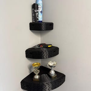 May include: Three-tiered corner shelves made from black tires. The top shelf holds a can of air freshener and a black spray bottle. The middle shelf has tools, and the bottom shelf displays two trophies.
