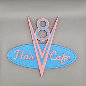 May include: A retro-style sign for Fla's Cafe, featuring a pink V8 emblem with a blue outline above a blue oval with the cafe's name in pink script. The sign has a vintage aesthetic.
