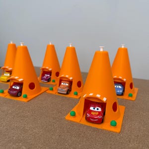 May include: Orange traffic cone-shaped toy structures with small toy cars inside. The cones have a square base with green spheres at the corners. The toy cars are various colors, including red, orange, and blue.