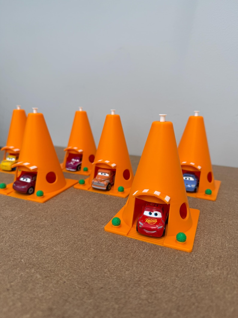 May include: Orange traffic cone-shaped toy structures with small toy cars inside. The cones have a square base with green spheres at the corners. The toy cars are various colors, including red, orange, and blue.