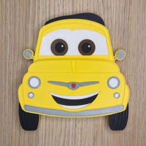 May include: A yellow cartoon car-shaped object with a smiling face, large brown eyes, and black tires. The car has a black roof and silver accents on the front bumper and around the headlights. The design is reminiscent of a classic car.