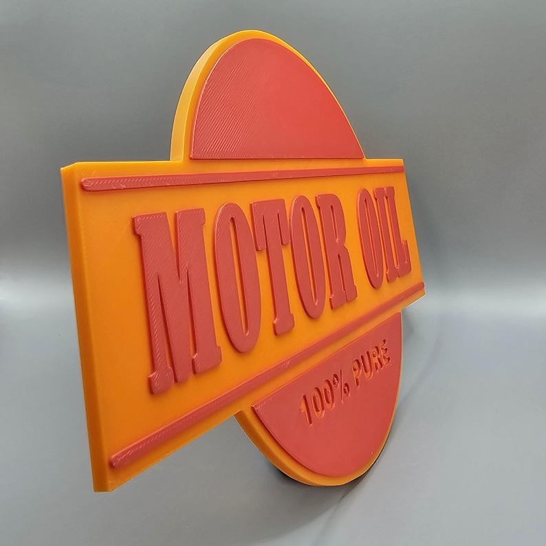 Puede incluir: Letrero naranja y rojo con las palabras "MOTOR OIL" en letras en relieve. El letrero tambi&eacute;n incluye el texto "100% PURE". El letrero tiene un dise&ntilde;o en capas con una parte superior e inferior redondeadas.