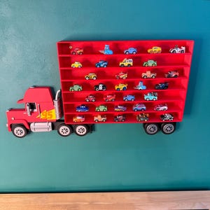 May include: A red wall-mounted display shelf shaped like a semi-truck with a trailer featuring multiple horizontal rows. The truck cab has a yellow lightning bolt graphic with the number 95. The trailer contains numerous small toy cars.