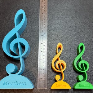 Three personalised treble clef ornaments in blue, green, and yellow, engraved with names. A ruler shows size options, with the largest around 20 cm tall. Perfect as a musician gift, music teacher decoration, or desktop ornament for music lovers.