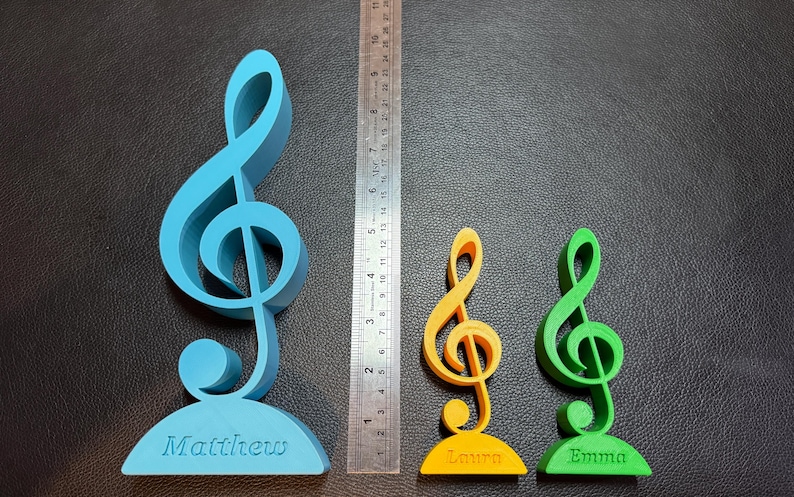Three personalised treble clef ornaments in blue, green, and yellow, engraved with names. A ruler shows size options, with the largest around 20 cm tall. Perfect as a musician gift, music teacher decoration, or desktop ornament for music lovers.