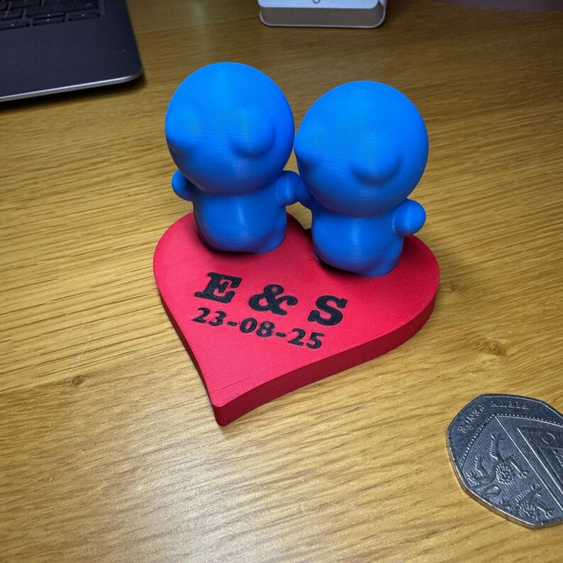 Cute 3D-printed cake topper with two blue figurines on a red heart base, engraved with initials and a date. Perfect for weddings or anniversaries, this cute eco-friendly PLA decoration is fully customisable and ideal for any special event.