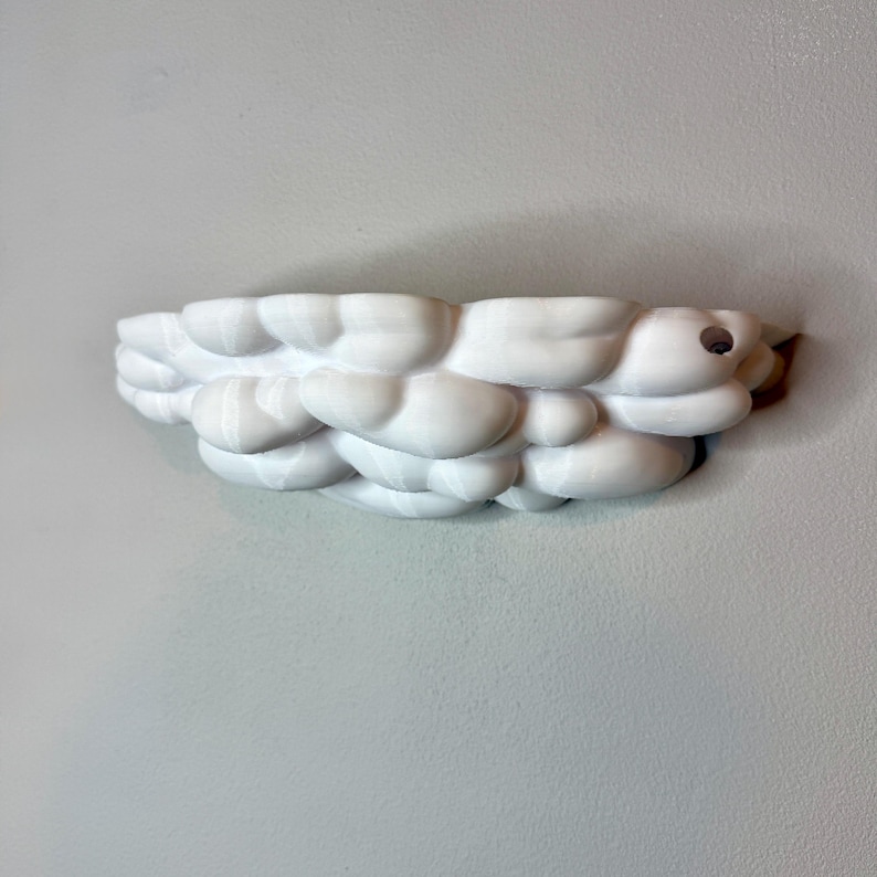 May include: A white, cloud-shaped wall shelf. The shelf is made of a series of rounded, puffy forms, resembling clouds. It is mounted on a light gray wall. The shelf is designed to hold small items and adds a whimsical touch to any room.