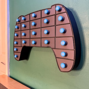 May include: A wall-mounted advent calendar shaped like a video game controller. It has a dark gray frame with 24 small drawers, each with a blue knob and a number from 1 to 24. The controller is mounted on a green wall.