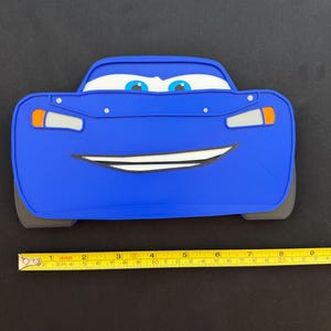 May include: A blue Lightning McQueen car-shaped object with a smiling face, white eyes, and orange and white headlights. The object is approximately 8 inches long. The car is on a black surface.