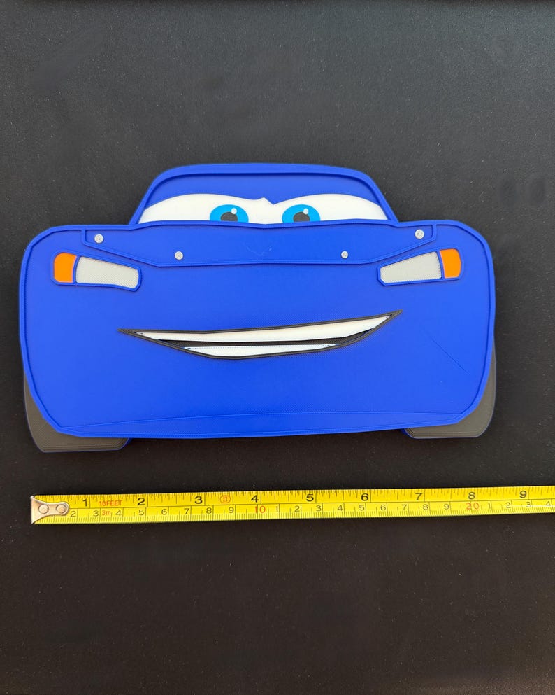 May include: A blue Lightning McQueen car-shaped object with a smiling face, white eyes, and orange and white headlights. The object is approximately 8 inches long. The car is on a black surface.