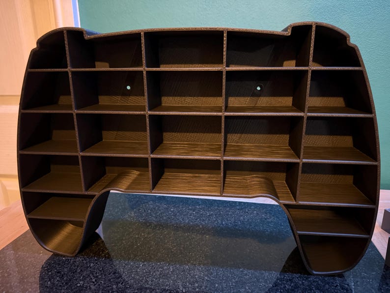 May include: A brown, multi-compartment shelf unit, resembling a video game controller. The unit has a series of small, square shelves, ideal for displaying small collectibles or organizing items. The overall design is modern and functional.