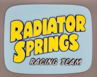 May include: A light blue sign with the words "RADIATOR SPRINGS" in yellow, outlined in black, and "RACING TEAM" in white. The sign has rounded corners and a light gray background.