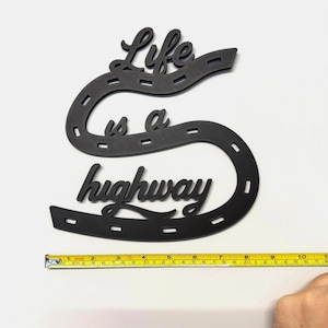 May include: Black wooden wall art in the shape of a large letter "S" with the words "Life is a highway" cut out. The art piece is approximately 11 inches long. The design resembles a road.