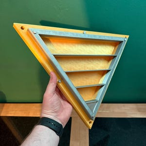 May include: A yellow and silver triangular shelf with three horizontal shelves. The shelf is held by a person's hand against a green background. The shelf has a silver frame and is secured with small silver screws.