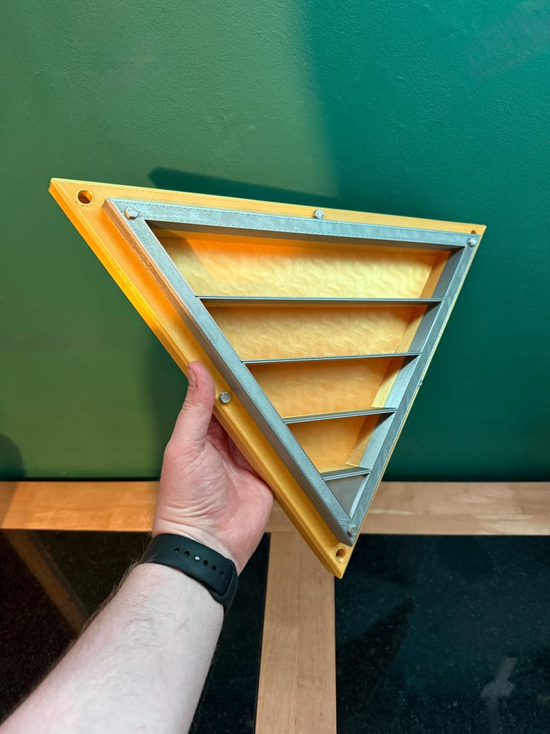 May include: A yellow and silver triangular shelf with three horizontal shelves. The shelf is held by a person's hand against a green background. The shelf has a silver frame and is secured with small silver screws.
