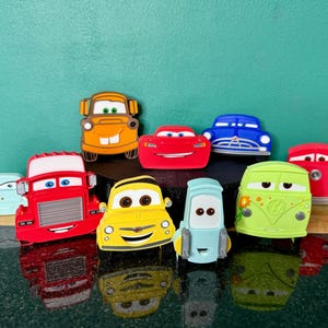 May include: A collection of colorful toy cars, including red, yellow, blue, and green vehicles. The cars have smiling faces and various designs, with a reflective surface below.