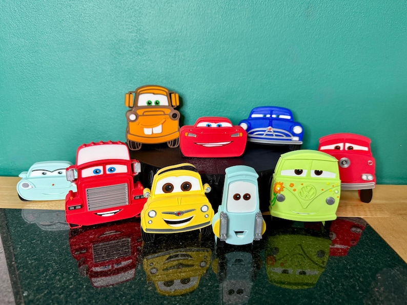 May include: A collection of colorful toy cars, including red, yellow, blue, and green vehicles. The cars have smiling faces and various designs, with a reflective surface below.