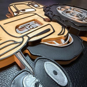 以下が含まれることがあります： ベージュ、黒、シルバーの色合いの3Dプリントされたおもちゃの車。ラジエーター、ヘッドライト、ホイールなどの詳細が特徴です。表現力豊かな目と笑顔の口を持つ、漫画のキャラクターを彷彿とさせるデザインです。