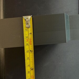 May include: A dark gray rectangular object with a matte finish, positioned next to a yellow measuring tape. The tape shows the object has a height of approximately 2.5 inches. The surface of the object displays fine horizontal lines.