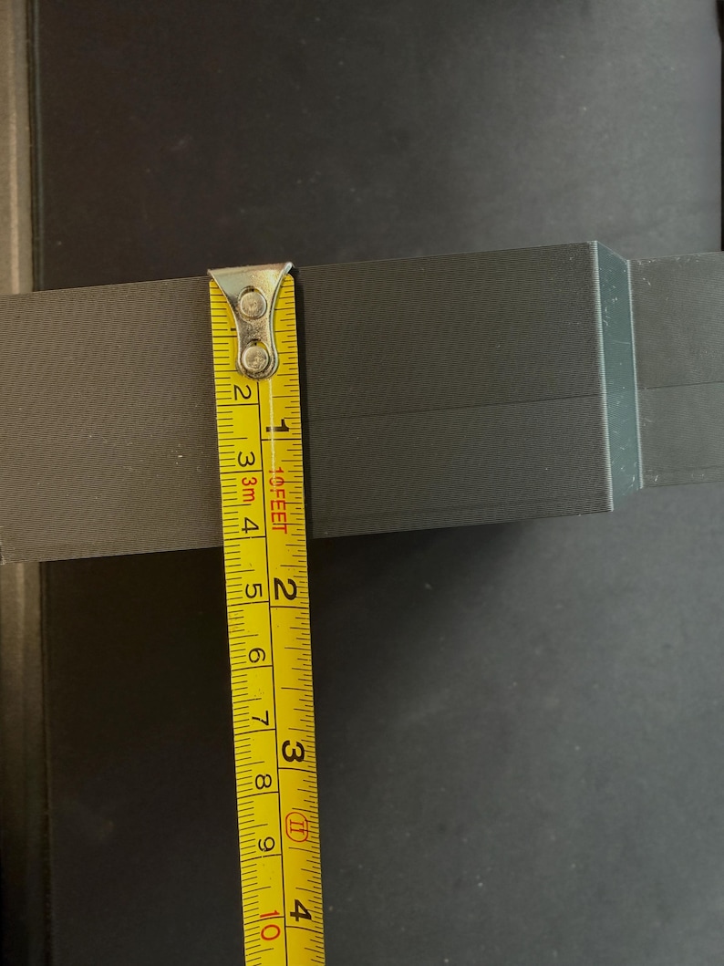 May include: A dark gray rectangular object with a matte finish, positioned next to a yellow measuring tape. The tape shows the object has a height of approximately 2.5 inches. The surface of the object displays fine horizontal lines.
