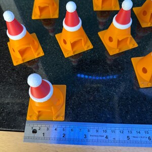 Może przedstawiać: Kolekcja pomarańczowych pachołk&oacute;w drogowych, każdy ozdobiony miniaturową czapką Świętego Mikołaja. Stożki mają około 5 cm wysokości i są ułożone na ciemnej powierzchni. Obok jednego stożka umieszczono linijkę dla skali.