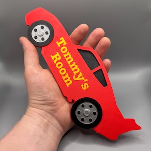 May include: Red car-shaped sign with the text "Tommy's Room" in yellow. The sign has black windows and gray wheels. The sign is held in a hand.
