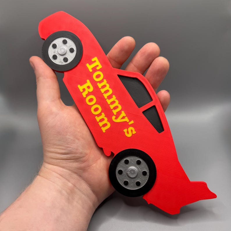 May include: Red car-shaped sign with the text "Tommy's Room" in yellow. The sign has black windows and gray wheels. The sign is held in a hand.