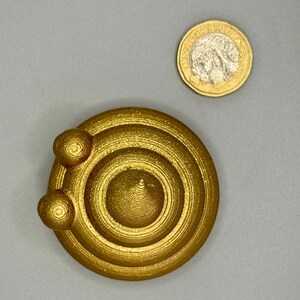 Top-down view of a 5cm diameter gold glittery 3D-printed poop emoji figurine next to a one-pound coin for scale. The figurine is round with a swirled texture, made from recycled plastic and hand-finished.