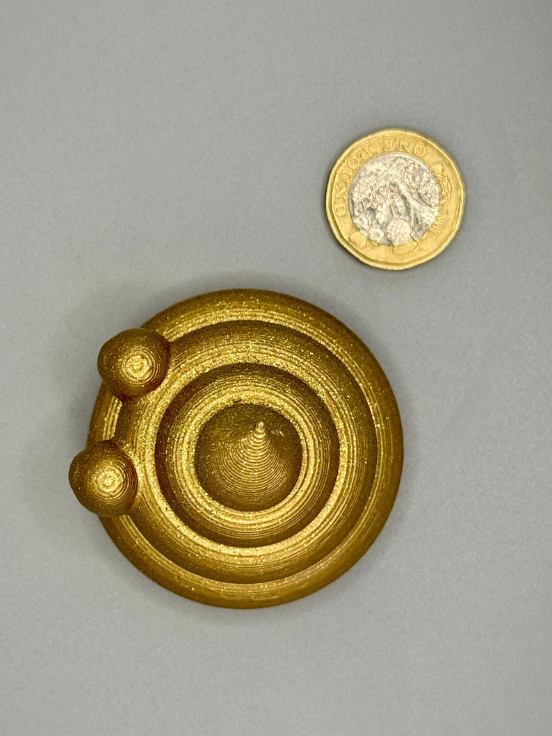 Top-down view of a 5cm diameter gold glittery 3D-printed poop emoji figurine next to a one-pound coin for scale. The figurine is round with a swirled texture, made from recycled plastic and hand-finished.