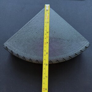 May include: A dark gray, triangular object with a textured surface, approximately 8 inches (20 cm) long, is measured with a yellow tape measure. The object appears to be a 3D-printed item, possibly a filter or a component.