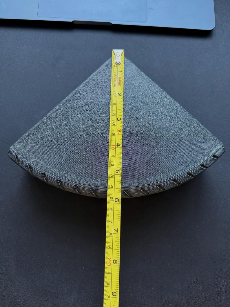 May include: A dark gray, triangular object with a textured surface, approximately 8 inches (20 cm) long, is measured with a yellow tape measure. The object appears to be a 3D-printed item, possibly a filter or a component.