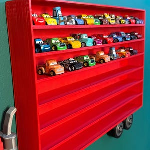 May include: A bright red rectangular wall-mounted display shelf with multiple horizontal tiers, currently holding a collection of small colorful toy cars. The shelf features a structured design with visible layered textures and two circular wheels at the base.