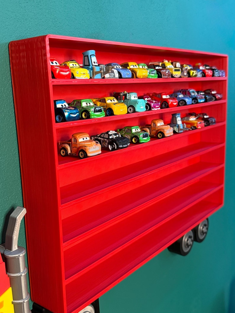 May include: A bright red rectangular wall-mounted display shelf with multiple horizontal tiers, currently holding a collection of small colorful toy cars. The shelf features a structured design with visible layered textures and two circular wheels at the base.
