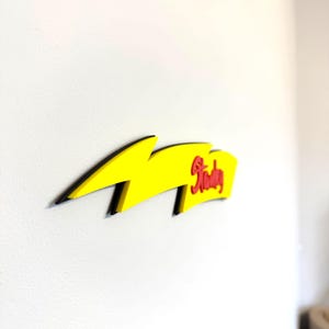 May include: A yellow lightning bolt-shaped wall decoration with black trim. The word "Stanley" is written in red script on the right side of the bolt. The decoration is mounted on a white wall.