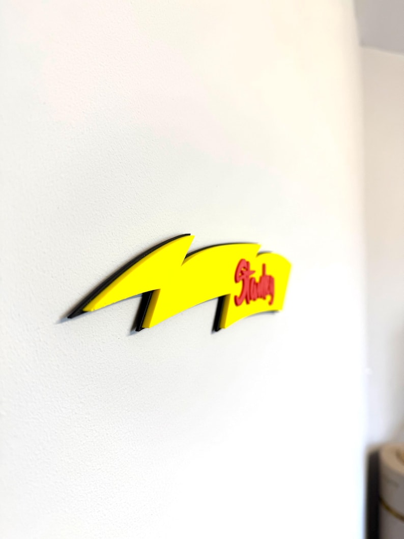 May include: A yellow lightning bolt-shaped wall decoration with black trim. The word "Stanley" is written in red script on the right side of the bolt. The decoration is mounted on a white wall.