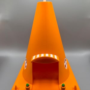 Op de afbeelding: Een 3D-geprint oranje kegelvormig huis met een wit zonnescherm boven de deur. Het huis heeft twee kleine ramen en een witte antenne op het dak. Er staan twee kleine groene planten in oranje potten aan weerszijden van de deur.