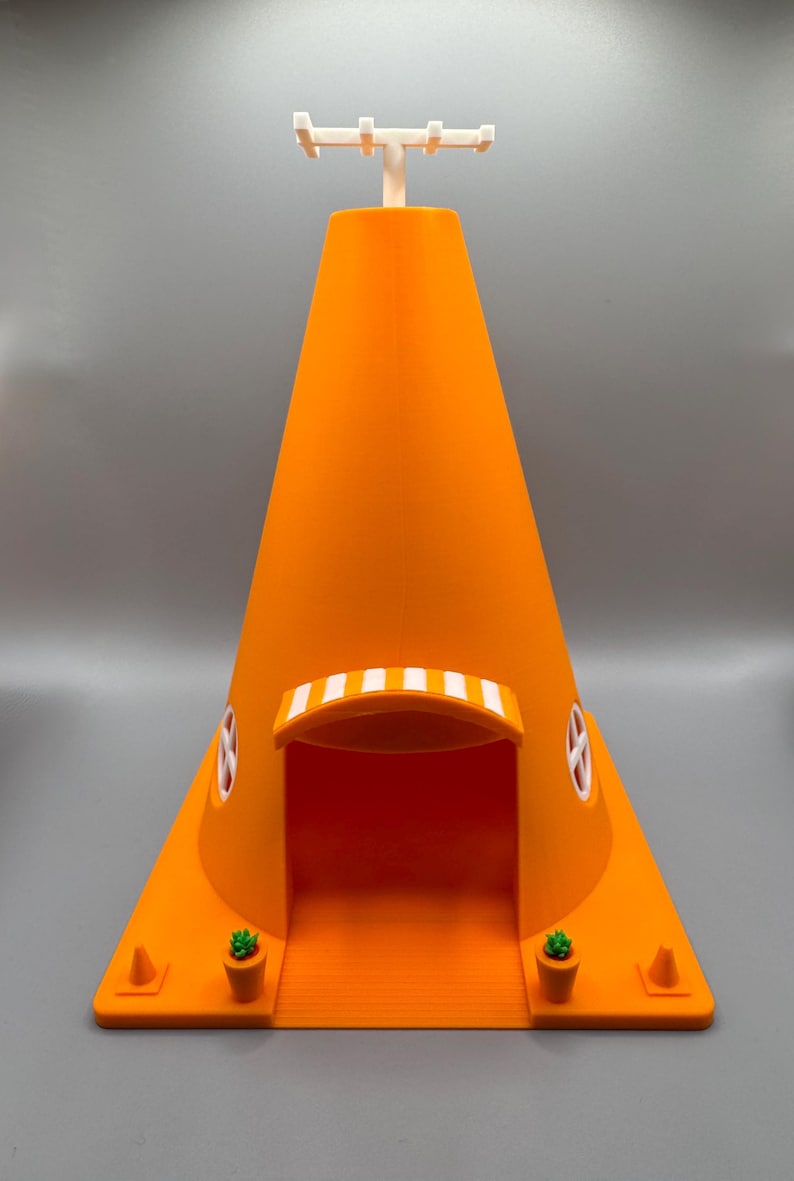 Op de afbeelding: Een 3D-geprint oranje kegelvormig huis met een wit zonnescherm boven de deur. Het huis heeft twee kleine ramen en een witte antenne op het dak. Er staan twee kleine groene planten in oranje potten aan weerszijden van de deur.