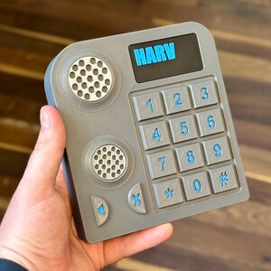 May include: A gray, rectangular device with a keypad and speaker holes. The keypad has blue numbers and symbols. The word "HARV" is displayed in blue at the top. The device is held in a hand.