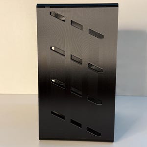 May include: A black, rectangular display stand with a series of angled, rectangular cutouts. The stand is made of a matte black material and is designed to hold and showcase items. The cutouts are evenly spaced and add a modern aesthetic.