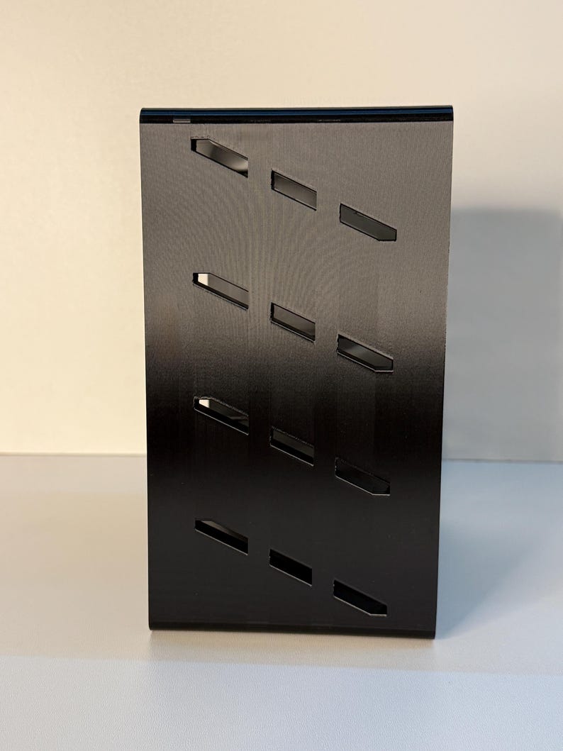 May include: A black, rectangular display stand with a series of angled, rectangular cutouts. The stand is made of a matte black material and is designed to hold and showcase items. The cutouts are evenly spaced and add a modern aesthetic.