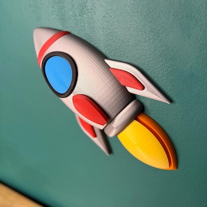 Può includere: Un razzo stampato in 3D nei toni del grigio, rosso, blu e giallo. Il razzo ha una striscia rossa attorno alla parte superiore, alette rosse e una fiamma gialla. L'oblò del razzo è blu. Il razzo è montato su una superficie verde acqua.