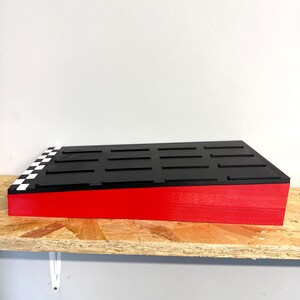 May include: A rectangular, tiered display stand with a red base and a black top. The top features evenly spaced black bars and a checkered flag design on one corner. The stand is on a wooden shelf.