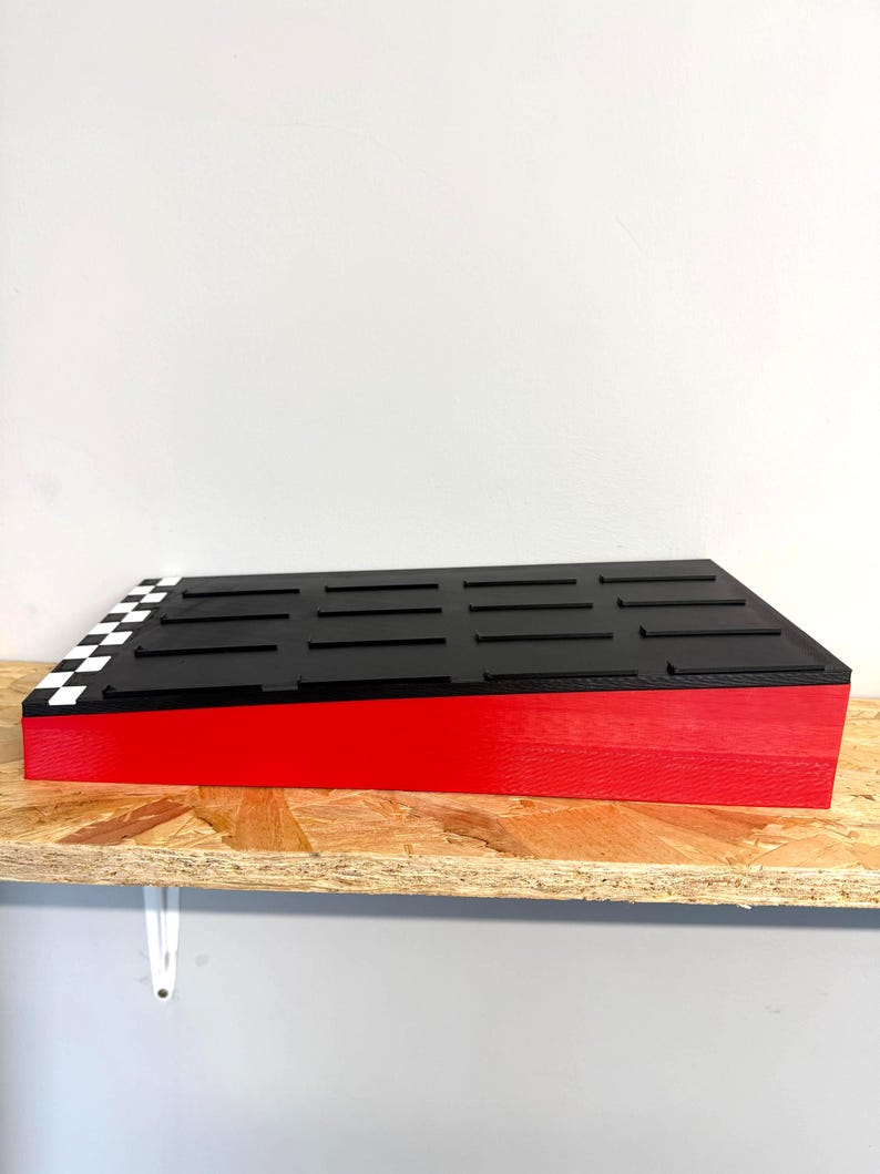 May include: A rectangular, tiered display stand with a red base and a black top. The top features evenly spaced black bars and a checkered flag design on one corner. The stand is on a wooden shelf.