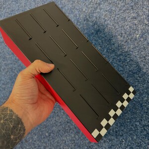 May include: A black and red rectangular box with a checkered pattern on one end. The top surface is black with evenly spaced, thin, raised lines. The box is held in a hand.